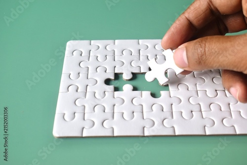Photography A close-up, angled view of a hand holding a white jigsaw puzzle piece directly over an open slot, demonstrating the concept of finding the perfect fit, strategic alignment, and problem-solving