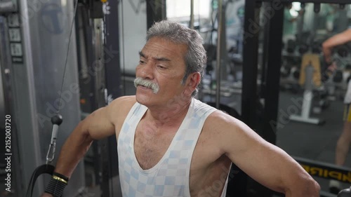 An Indian Asian fit, muscular elderly or old man performs a standing cable chest fly exercise during a resistance training session in a modern gym. Wellness, fitness, weight training workout concept
