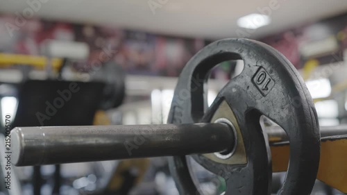 A close up shot of a weight plate is added to a barbell before performing resistance weight training or a workout, with a modern gymnasium visible in the background. Fitness, physical exercise concept