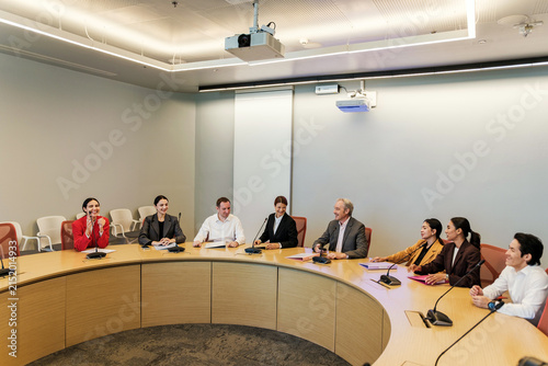 Group of professionals in a modern conference room holding a roundtable meeting with microphones and a projector screen, discussing plans and strategy.