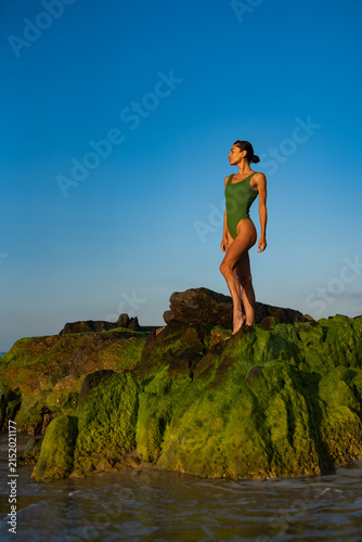 Confident woman in green swimsuit standing barefoot on mossy rocks by the ocean, basking in golden sunrise light, looking serene and strong with waves and blue sky in background.