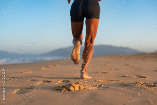 Back view close-up of athletic woman jogging barefoot on beach at sunrise, sand flying from her foot with every step, warm morning light and soft mountain horizon in background.
