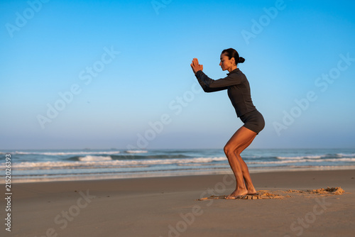фотографія Fit woman in sportswear doing squat exercise on sandy beach at sunrise, focusing on strength and posture, with calm ocean waves in background and clear blue morning sky