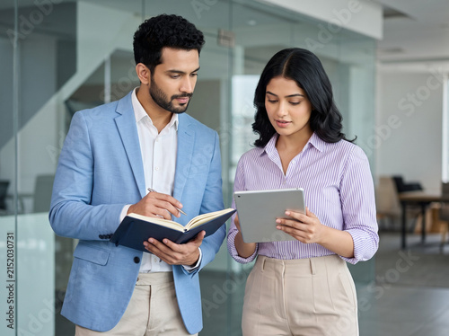 young Indian businesswoman using a tablet while a male professional takes notes in an open diary during a corporate meeting representing consulting teamwork and planning