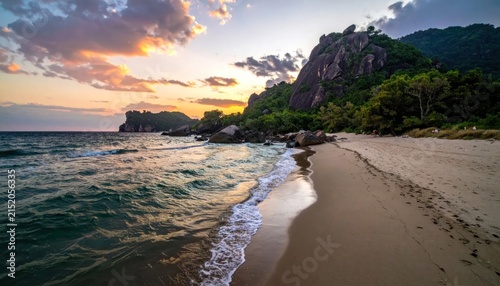 Idyllic tropical beach at sunset with dramatic sky, lush green mountains, and calm ocean waves gently lapping the shore.