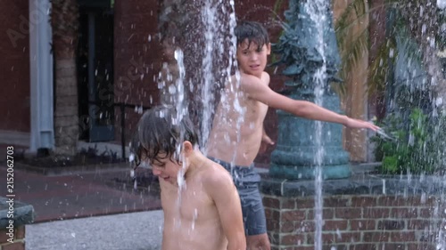 Two boys enjoy cooling off beneath the water jets of an outdoor splash fountain in a residential setting. The candid scene captures carefree childhood, active play.