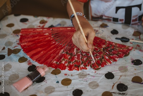 Hand meticulously paints a vibrant red fan using a brush. The scene captures the creative process on a polka dot-covered table, highlighting artistic expression and focus.