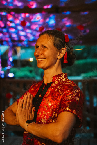 A woman in a traditional red Chinese dress stands smiling, clasping her hands in greeting or gratitude. The attractive background creates a festive atmosphere, highlighting her joyful expression.