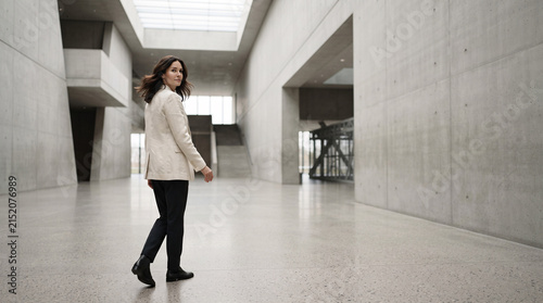 A confident woman walking through a modern minimalist concrete building hall.