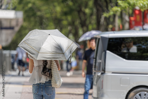 Young woman in a white ruffled blouse and jeans holding a UV umbrella on a sunlit city street with blurred traffic and pedestrians.