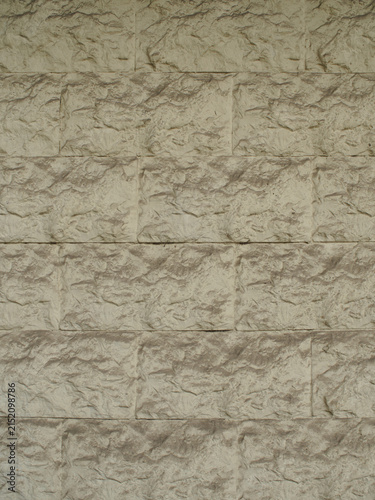 A vertical photograph of a light beige decorative stone block wall with a rough, embossed texture, background with subtle highlights and shadows of wall