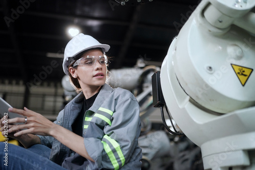 Female engineer in safety gear inspects robotic arm with tablet in modern factory