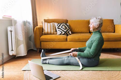 Senior woman training at home, sitting on green mat in fitness clothes and using rubber resistance band, following online video tutorial on laptop, doing physical exercise for strong muscles and bones