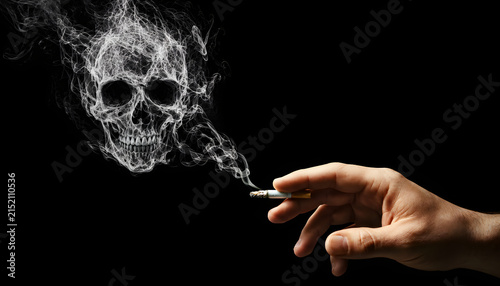 A hand holds a burning cigarette with smoke artistically forming the shape of a human skull against a dark black background, symbolizing the dangers of tobacco.