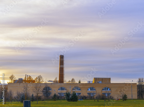 Cuadro en lienzo Tall brick industrial building with smokestack rising under soft pastel sunrise
