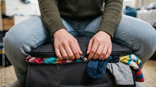 Close-up of unrecognizable young woman sitting on overcrowded suitcase, stress during travel, problems with packing luggage and difficulties with vacation planning