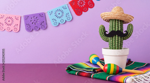 Festive Cinco de Mayo concept featuring a potted cactus wearing a sombrero and mustache, colorful maracas, a serape blanket, and papel picado banners against a purple background.