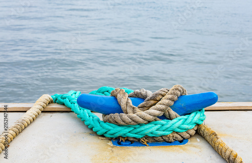 Mooring cleat painted blue with turquoise and hemp ropes on concrete pier against blurred sea water copy space