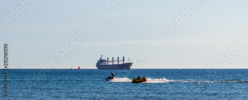  jet ski towing yellow inflatable tube under clear sky. large cargo ship with deck cranes with a damaged wheelhouse as a result of a drone attack, sailboats visible on horizon