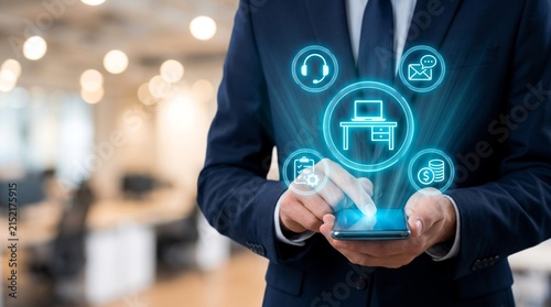 Professional businessman in dark suit touches glowing digital interface with business office icons floating above his modern mobile smartphone device