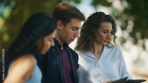 Three young colleagues engage in an outdoor discussion, reviewing documents and a tablet. They collaborate on a project, focused and concentrated in a green environment.