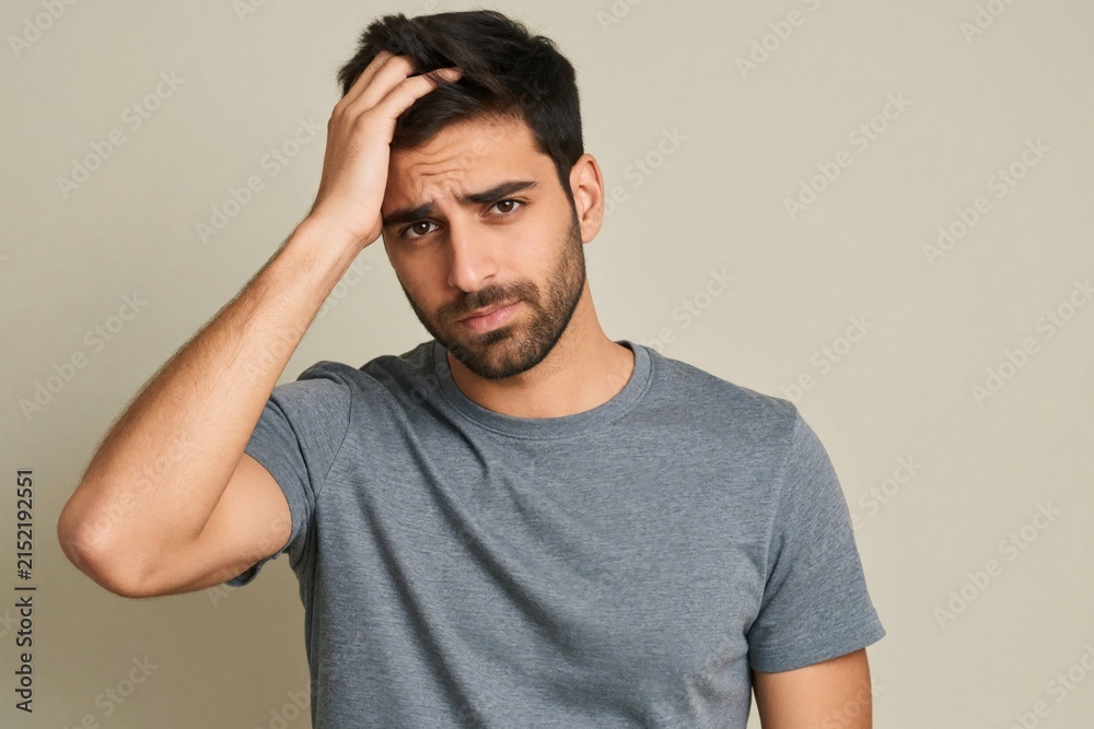 Fototapeta premium Young man with hand on head looks stressed. He shows clear signs of worry or deep thought. His facial expression conveys concern and unease.