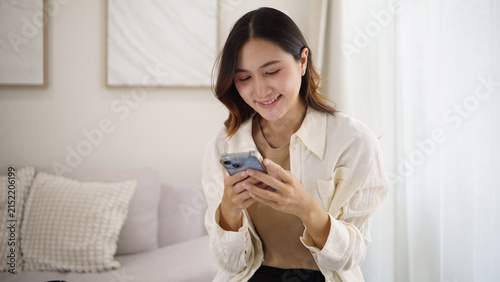 Young Asian woman using a smartphone while relaxing with coffee in her living room.