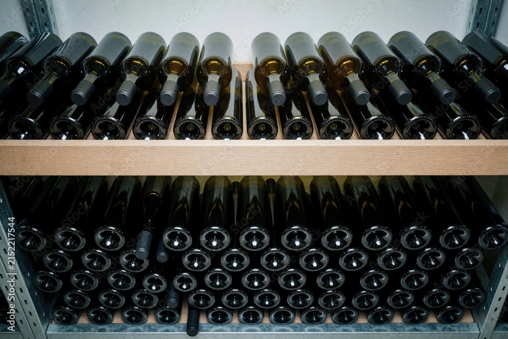 Obraz premium Many dark glass bottles for wine are neatly arranged on wooden shelves in cellar. Bottles stored for aging and maturation process. Collection ready for tasting or sale.