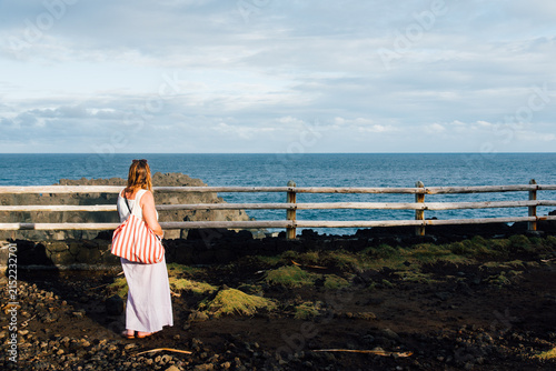 Femme regardant la mer. Vacancière contemplant l'horizon le soir au bord l'Océan Indien. Seule face à la mer. Touriste avec un sac rouge à La Réunion
