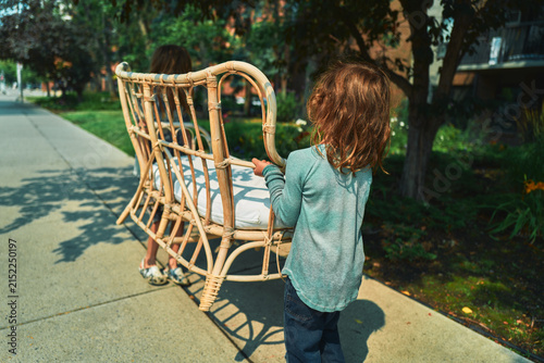 Two boys carrying a wicker loveseat down a sidewalk from behind