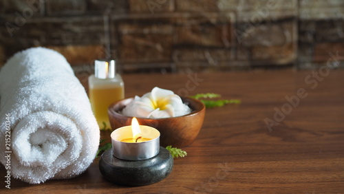 Soothing spa and wellness composition featuring a clean rolled white towel, a lit tealight candle on a dark basalt stone, a frangipani plumeria flower with bath salts in a wooden bowl, and an essentia