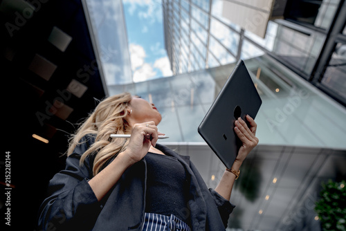 Obraz na plátně In profile beneath the glass atrium, an Asian businesswoman lifts her chin high, stylus resting at her lips as her tablet is held aloft beside her, beneath the office towers
