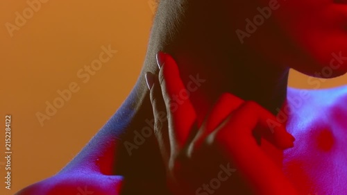 Tight crop on young woman lit with blue and red gel lighting touching her neck and face skin with two fingers on a solid orange background | Perfume campaign footage