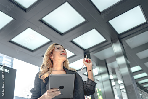 Quadro su tela Asian businesswoman with long blonde-dyed wavy hair raises a stylus pen near her face while gazing upward, tablet in hand beneath a glowing glass skylight canopy above her