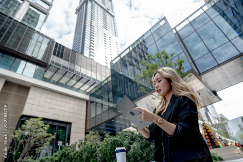 Photography Asian businesswoman walks past a modern office entrance lined with greenery, eyes fixed downward on her tablet screen while a takeaway coffee cup rests on a planter beside the path
