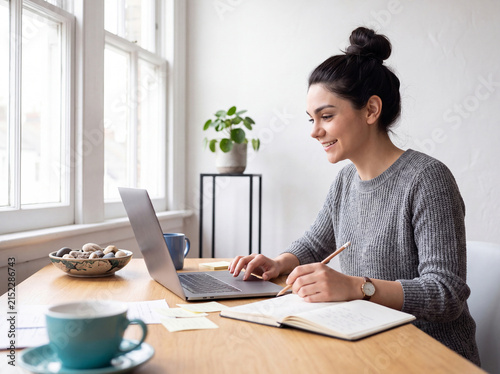 Focused young woman working remotely from her home office, typing on a laptop and taking notes in a journal.
