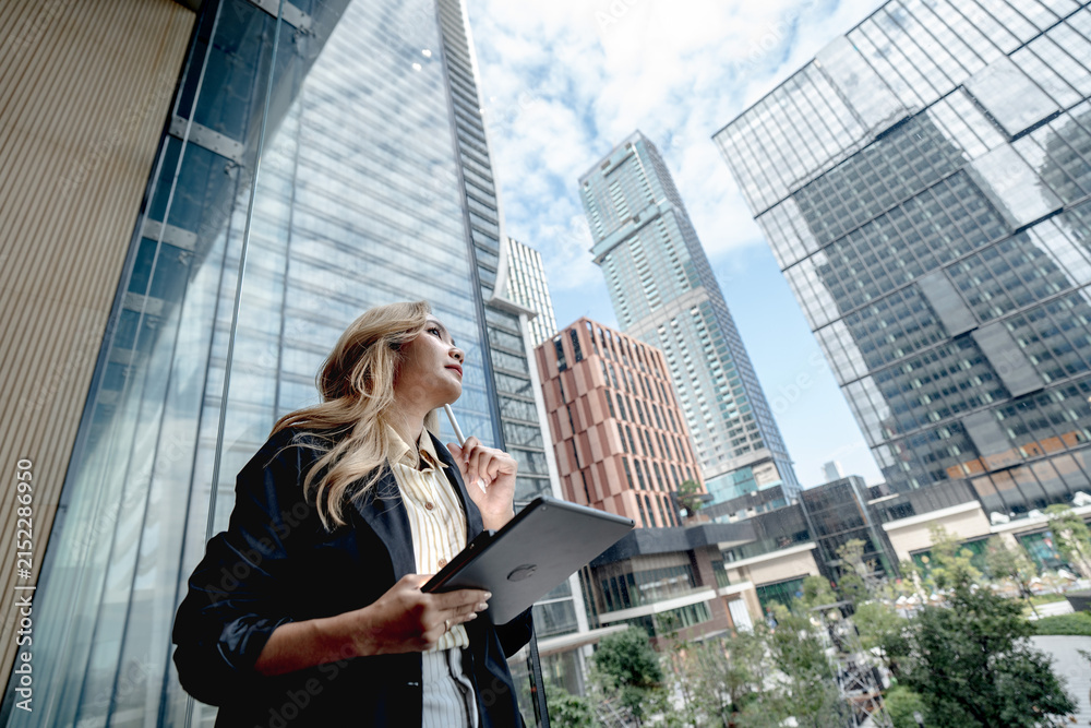 Obraz premium Asian businesswoman gazes upward pensively with a stylus near her lips, tablet held close to her chest while standing beneath soaring glass skyscrapers in Bangkok.