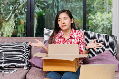 Disappointed Asian woman shrugging shoulders after opening parcel box at home. Sad customer receiving wrong product from online shopping.
