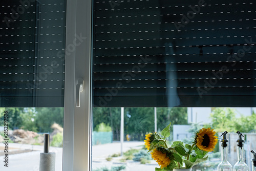 Partially closed roller shutters reduce daylight through a kitchen window, with sunflowers and glass bottles on the counter