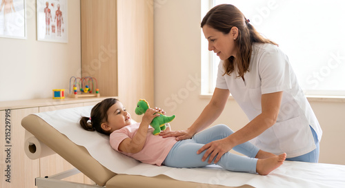 A smiling female pediatrician or physiotherapist examining the abdomen of a toddler girl holding a green dinosaur toy on a medical table.