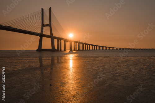 Golden sunrise over Vasco da Gama bridge and Tagus river reflecting on wet sand, Lisbon, Portugal