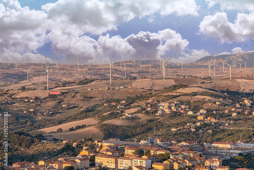 Wind turbines on rolling hills above a scenic rural town under a cloudy blue sky. Renewable energy landscape.