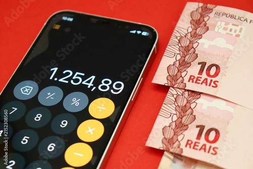 Smartphone displaying a calculator app next to Brazilian Real banknotes and coins. Brazilians financial planning and budgeting.