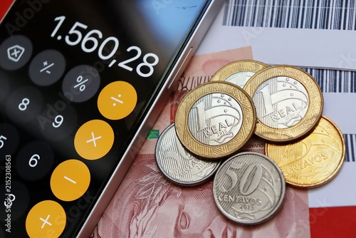 Smartphone displaying a calculator app next to Brazilian Real banknotes, payment slips and coins. Financial planning and budgeting. Brazilian money.