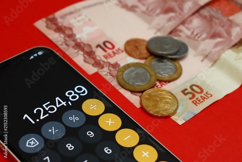 Smartphone displaying a calculator app next to Brazilian Real banknotes and coins. Brazilians financial planning and budgeting.