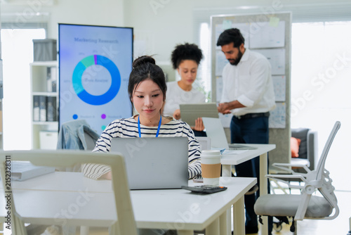 A focused employee works on her laptop in the foreground while colleagues collaborate at a desk behind her, capturing the layered activity of a busy, modern open-plan office in full swing.