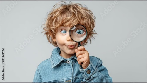 Young Caucasian boy with curly hair looking through a magnifying glass against a simple light grey studio background.