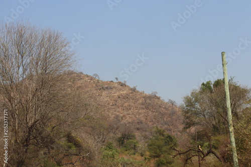 Scenic semi-arid mountainous landscape featuring rugged rocky hills, dry woodland, scattered trees, large natural boulders and a tranquil reservoir surrounded by rolling terrain. The collection captur