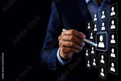 A businessman in a dark suit using a stylus to select a human from a digital grid of user profile symbols on a black background screen