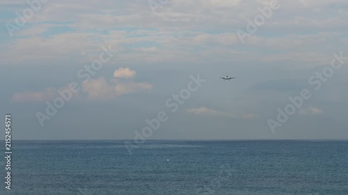 Passenger commercial airplane flying low over blue sea water approaching airport for landing under cloudy sky, wide angle static shot